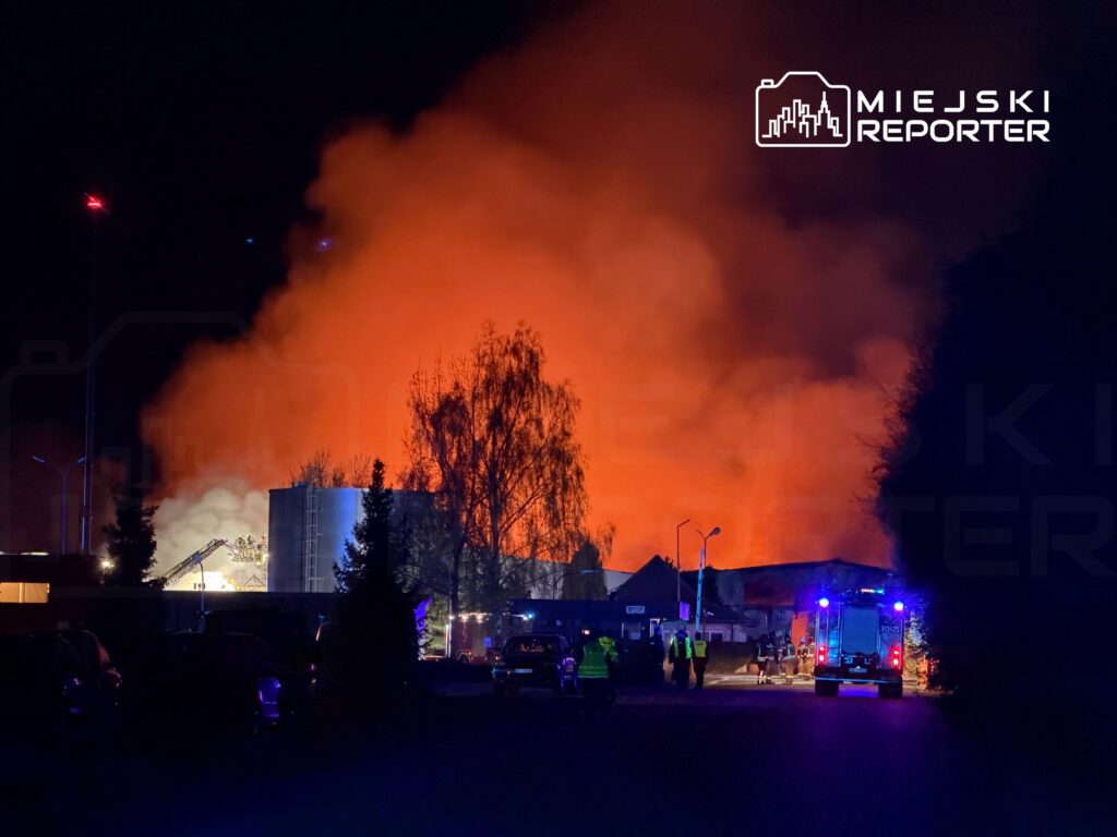 Ogromny pożar z intensywnym ogniem i dymem w nocy, w tle widać interwencję straży pożarnej i zaparkowane samochody.