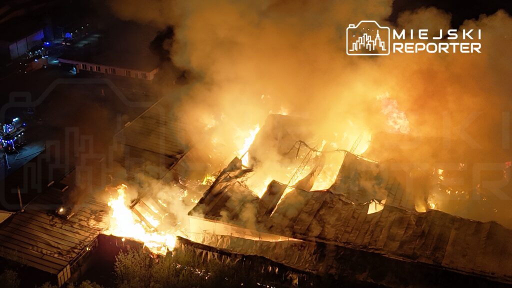 Wielki pożar z płomieniami i dymem unoszącym się nad zniszczonym budynkiem, w tle widać wozu strażackie.