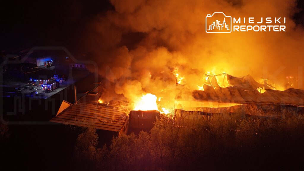 Ogień trawi dach budynku, a w tle widać pojazdy strażackie oraz dym unoszący się w nocy.