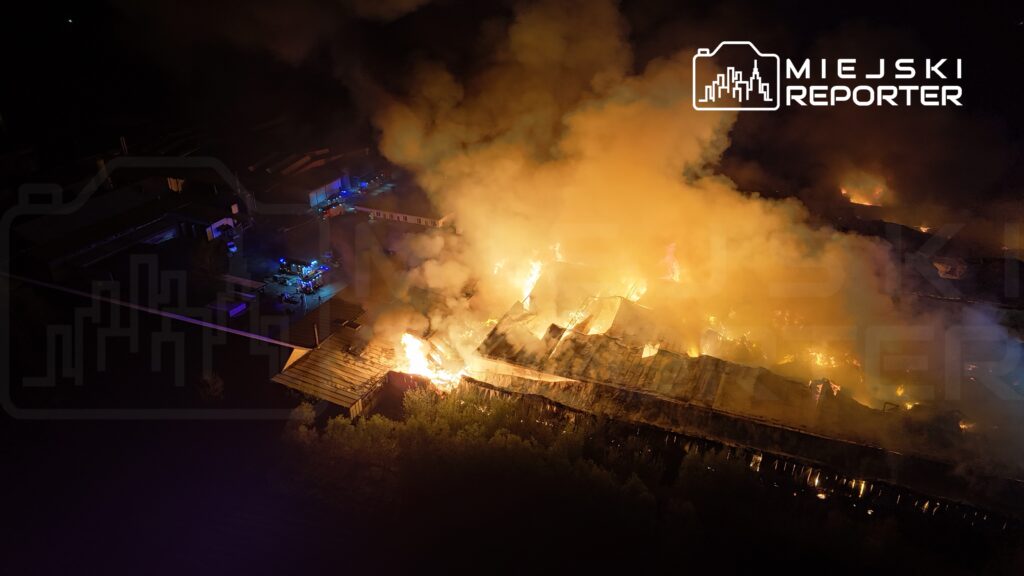 Intensywne płomienie i gęsty dym unoszą się nad zniszczonym budynkiem, w otoczeniu wozów strażackich i oświetlenia.