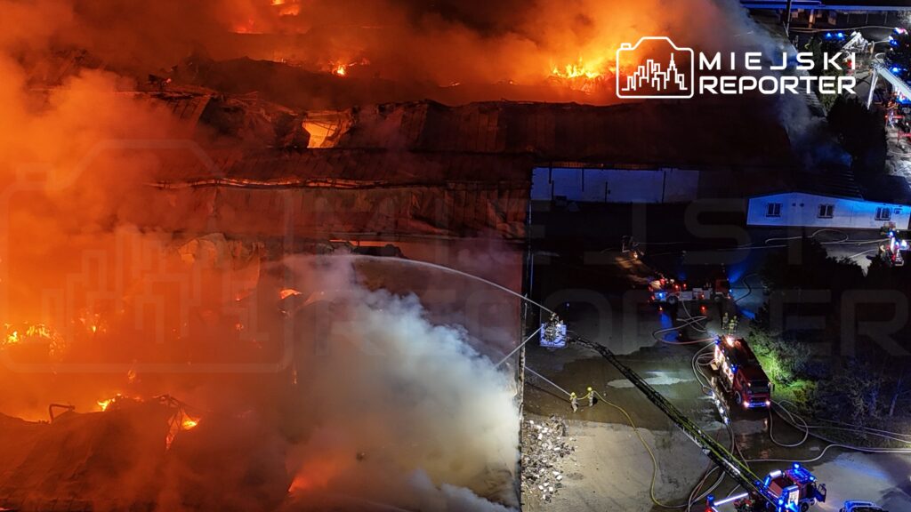 Pożar w dużym zakładzie przemysłowym, strażacy walczą z ogniem, używając węży gaśniczych w gęstym dymie.