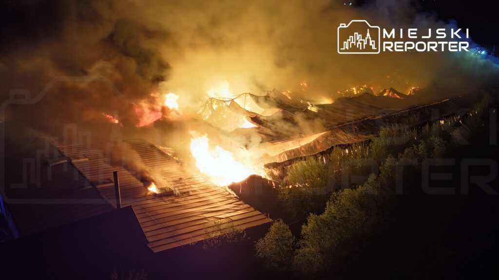Pożar strawił dach budynku, wydobywają się kłęby dymu i płomienie w nocy, w otoczeniu zieleni.