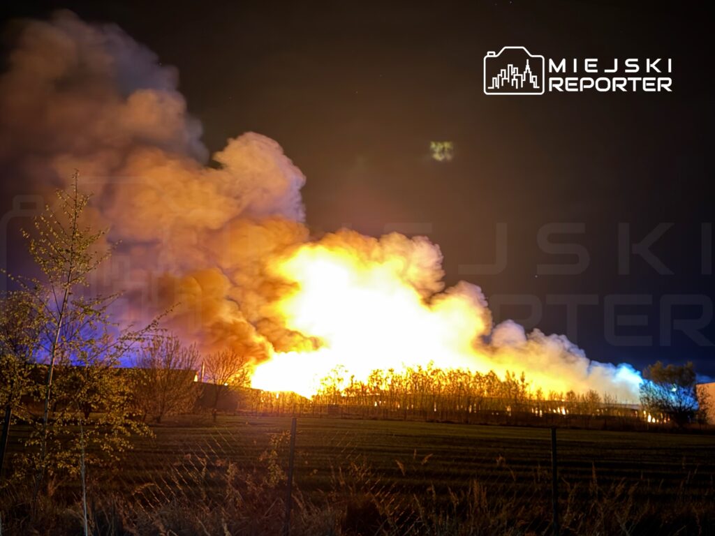 Ogromne płomienie i gęsty dym unoszą się nad terenem, otoczonym drzewami i ogrodzeniem.