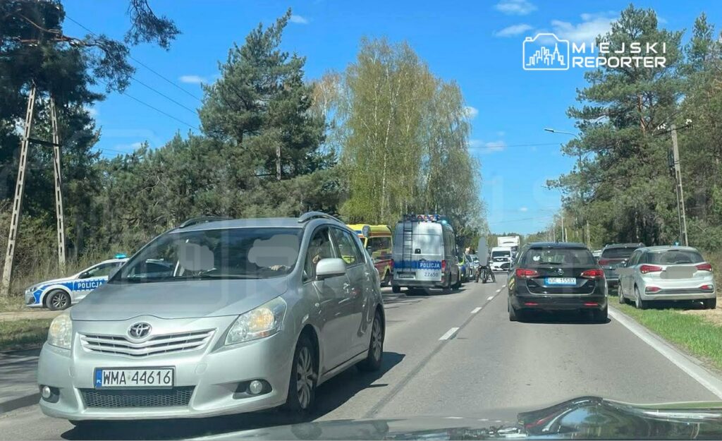 Ruch drogowy zatrzymany na drodze, w tle radiowozy policji oraz pojazdy służb ratunkowych.