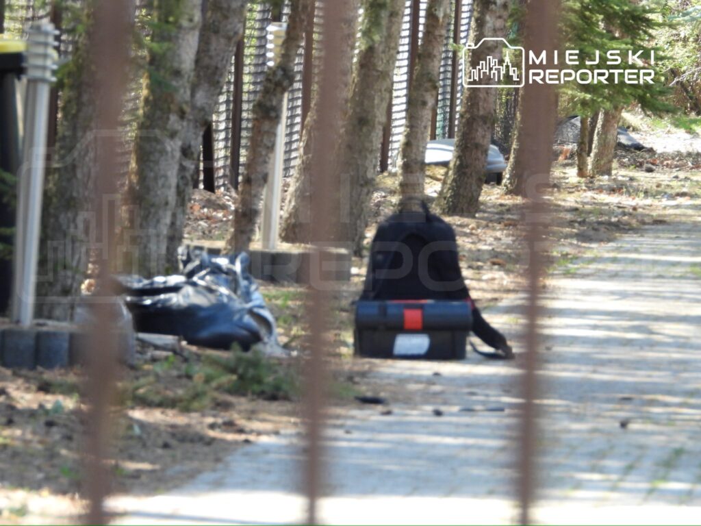 Osoba w czarnej odzieży siedząca na ścieżce obok torby i worka, w otoczeniu drzew i ogrodzenia.