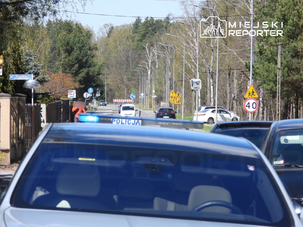 Policjant w niebieskiej koszuli stojący obok radiowozu na drodze, przy znaku ograniczenia prędkości do 40 km/h.