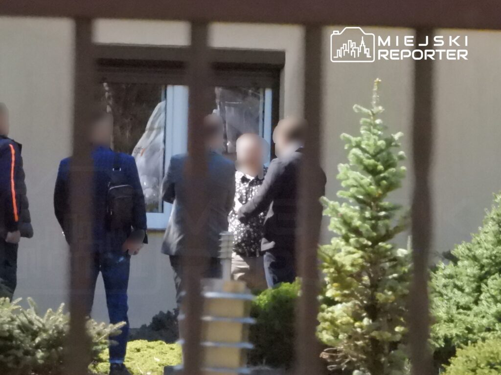 Grupa mężczyzn rozmawia na zewnątrz budynku, w tle widać okno z zasłonami. Wokół rosną krzewy.