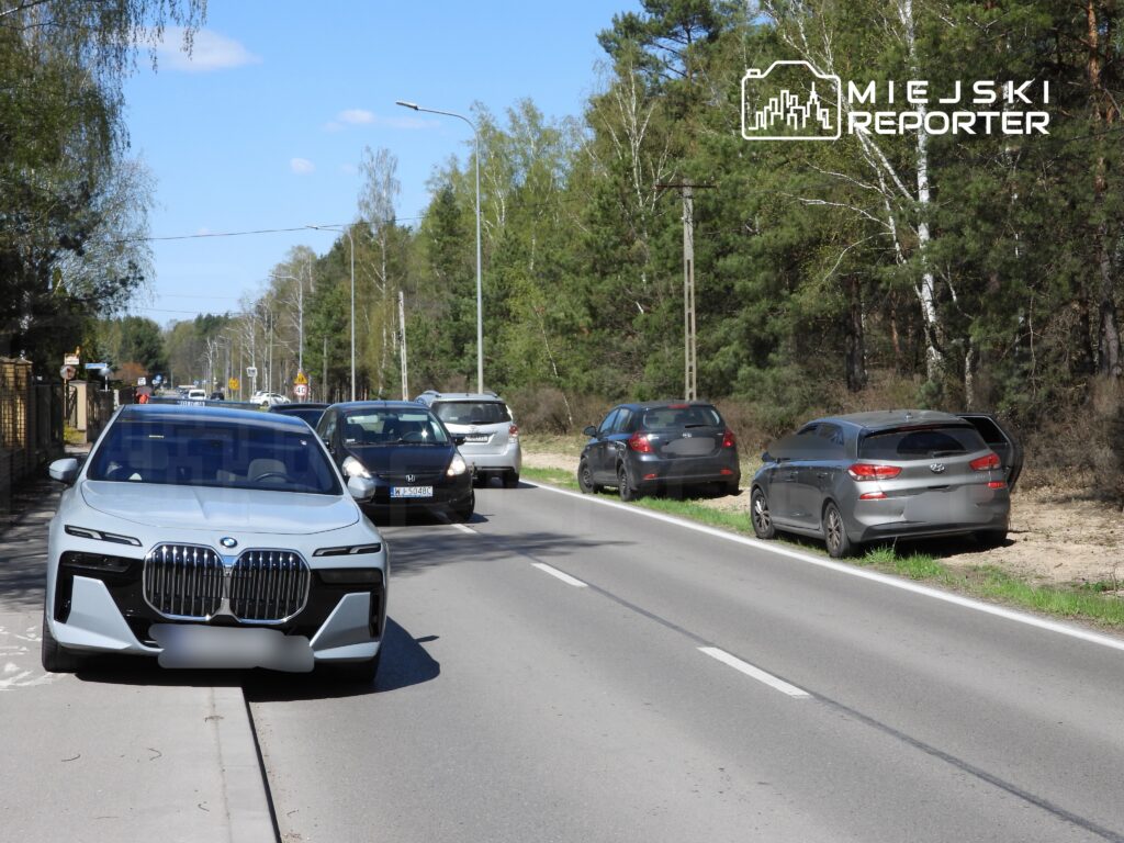 Jasnoszary samochód osobowy zaparkowany na poboczu drogi, obok ruchliwej jezdni z innymi pojazdami w tle.