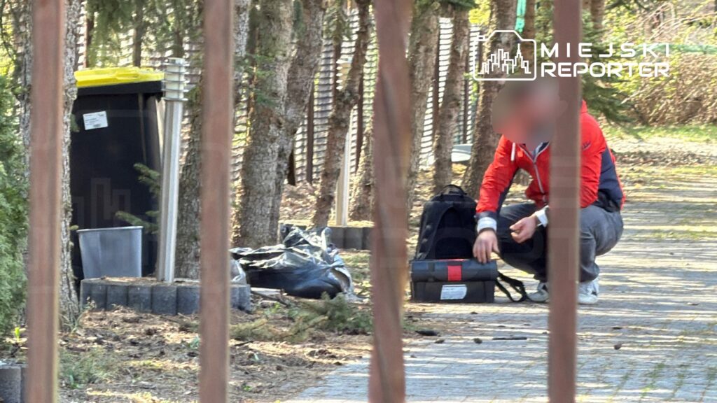 Mężczyzna w czerwonej bluzie siedzący na chodniku obok torby, przy śmietniku w otoczeniu drzew.