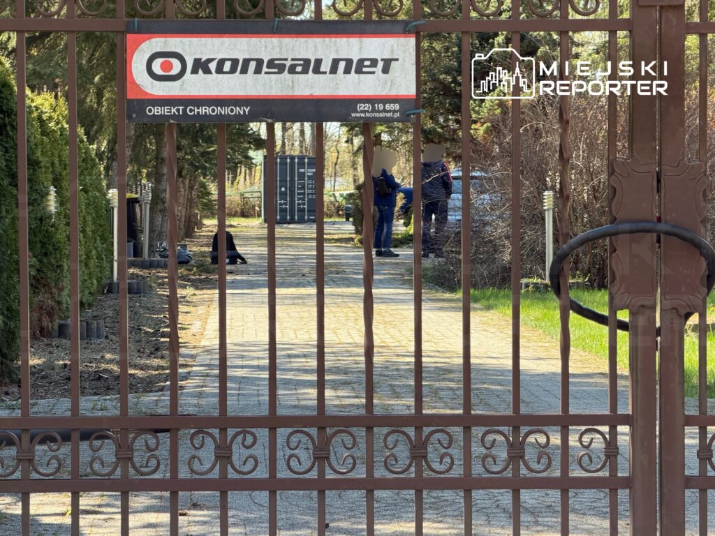 Na terenie otoczonym ogrodzeniem, pracownicy ochrony rozmawiają w pobliżu bramy z tablicą informacyjną.