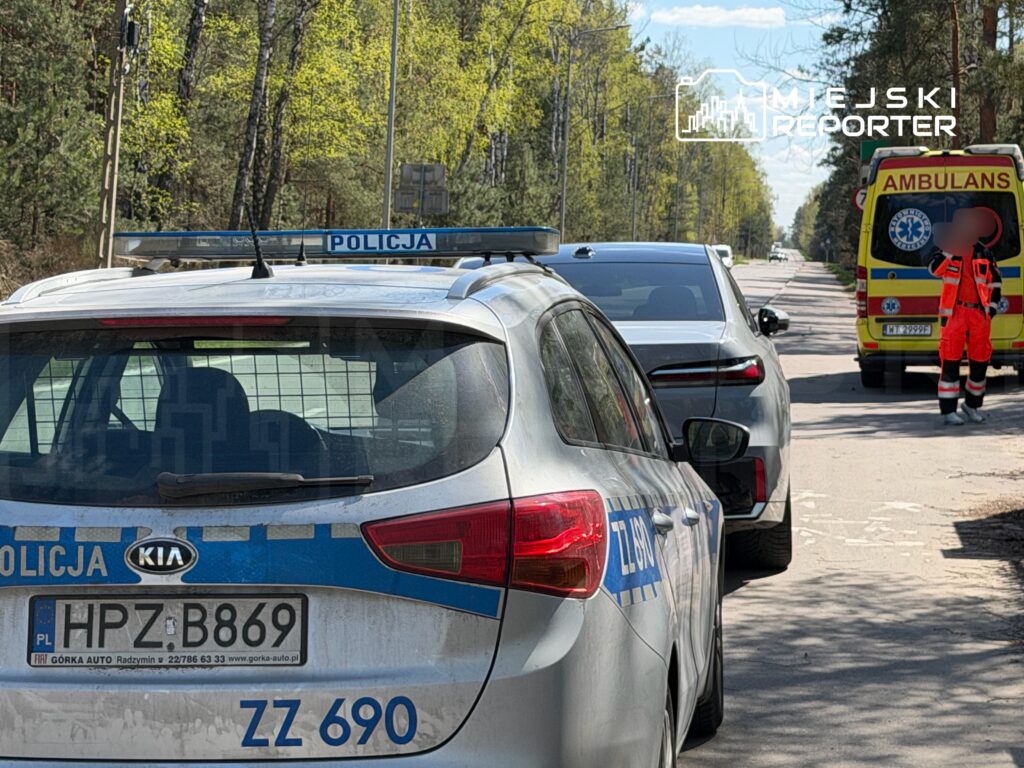 Policyjny radiowóz zatrzymany na drodze, obok karetka pogotowia oraz ratownik w pomarańczowym uniformie.