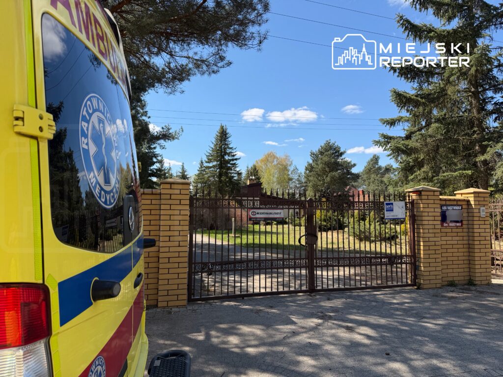 Ambulans z logo "Ratownictwo Medyczne" parkuje przy zamkniętej bramie otoczonej zielenią.