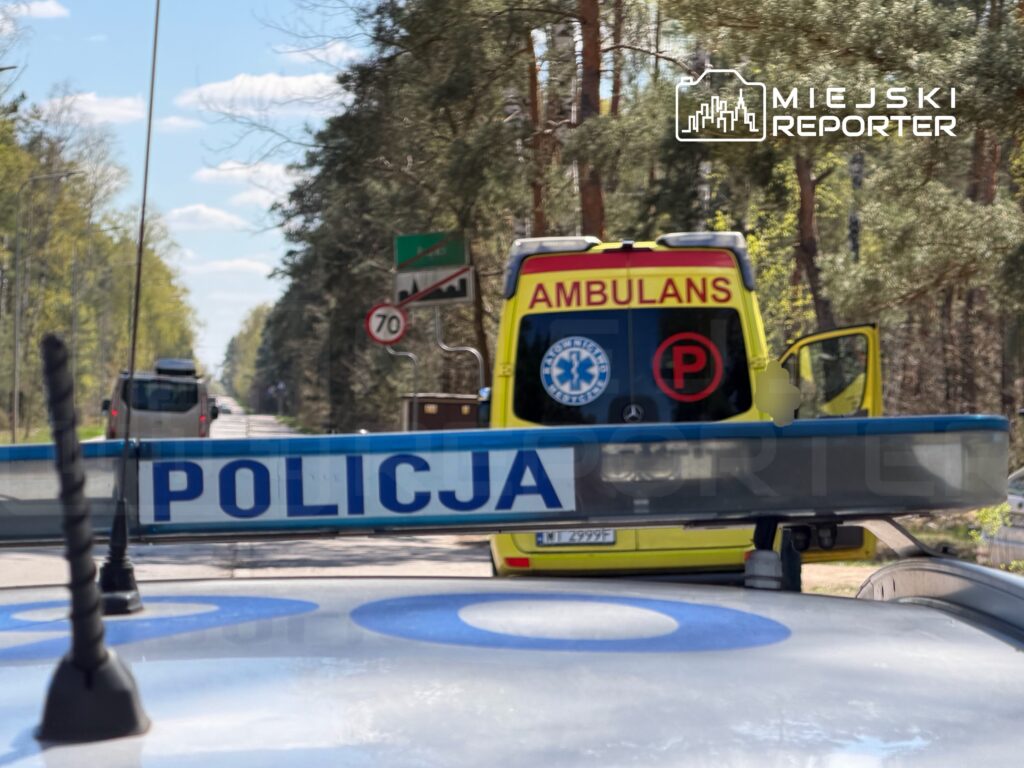 Policyjny radiowóz z napisem „POLICJA” na widoku, zatrzymujący ruch obok karetki pogotowia na leśnej drodze.