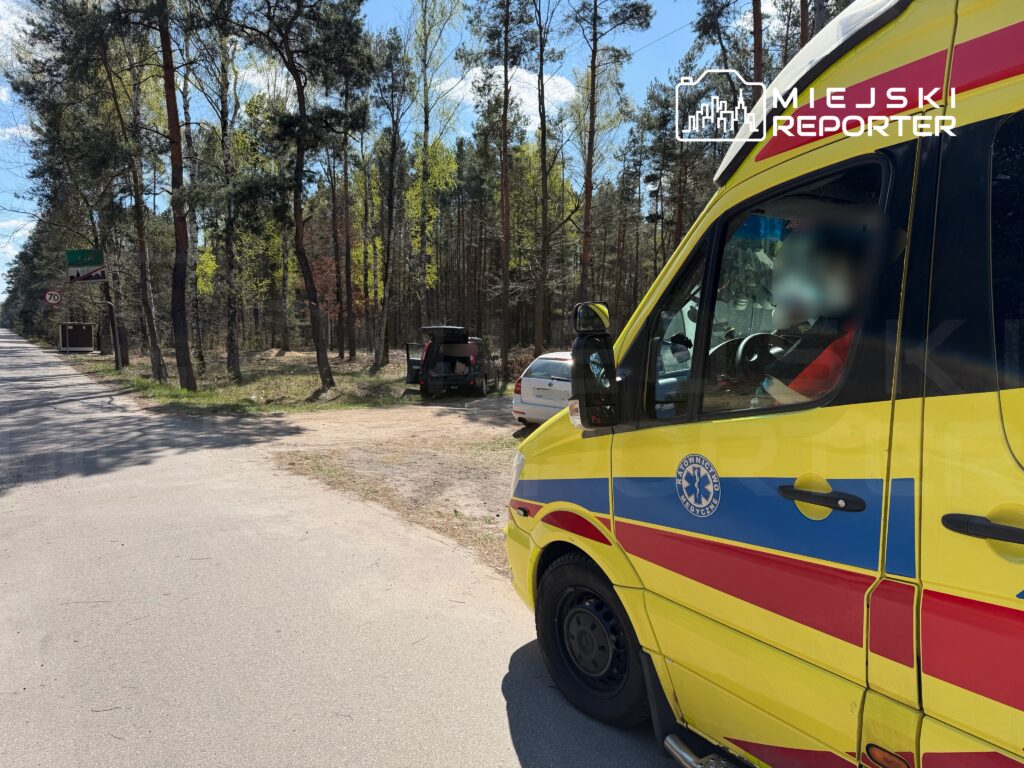 Karetka pogotowia stojąca w lesie obok drogi, obok niej zaparkowany samochód osobowy na poboczu.