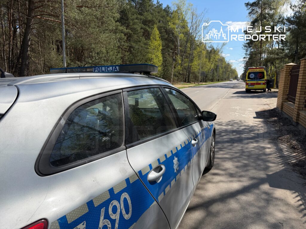 Radiowóz policyjny zatrzymany na poboczu drogi, obok stoi ambulans w lesistym otoczeniu.
