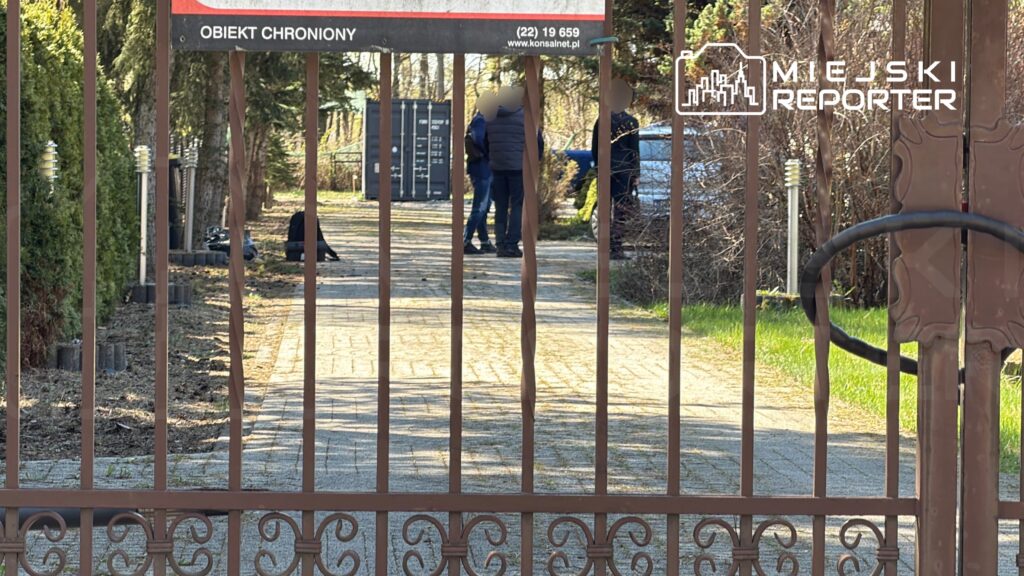 Grupa osób rozmawia na utwardzonej drodze przed bramą z napisem "OBIEKT CHRONIONY".