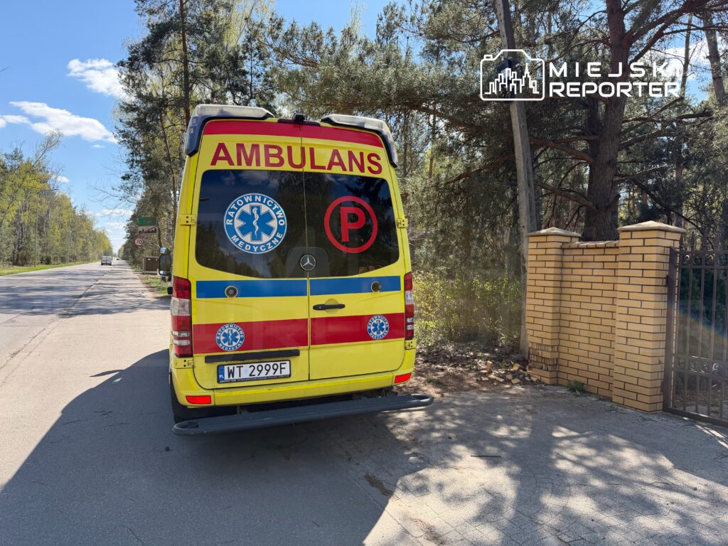 Ambulans z logo "Ratownictwo Medyczne" zaparkowany przy drodze, w otoczeniu drzew i ogrodzenia.