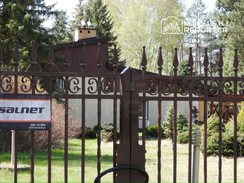 Metalowa brama z ozdobnymi detalami prowadzi do ogrodu z zielonymi krzewami i drzewami w tle.