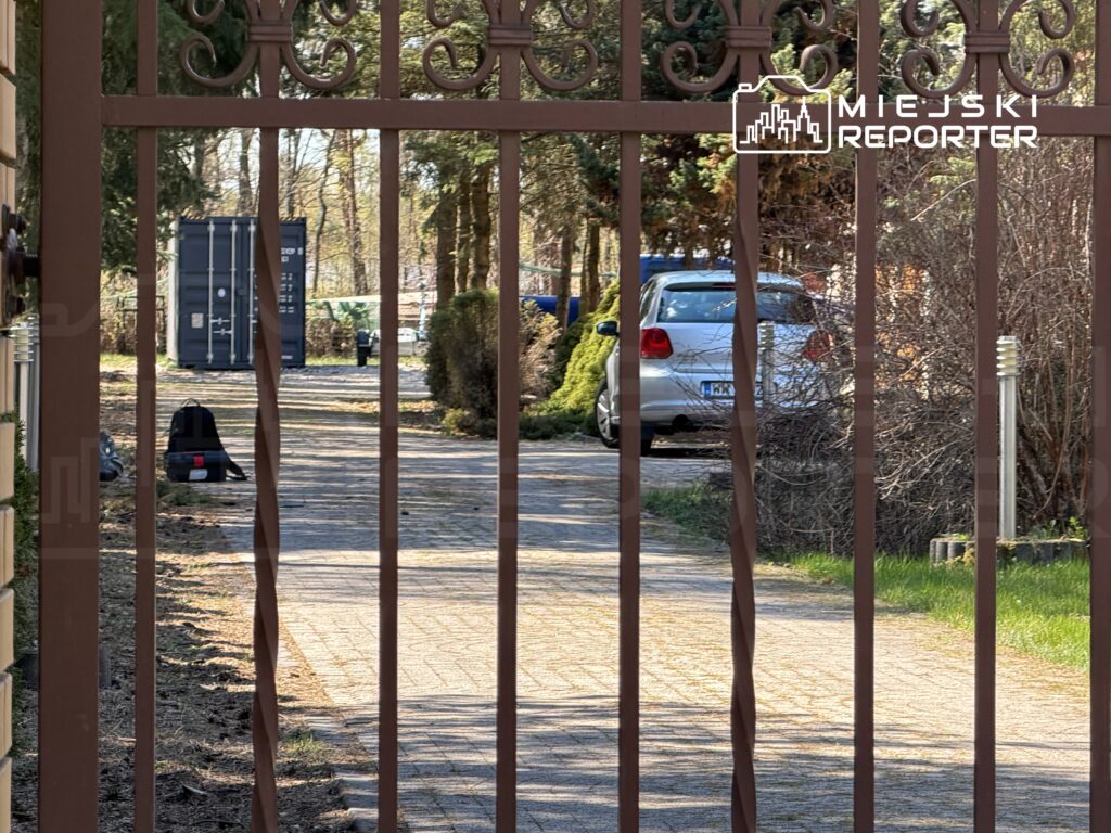 Zamknięta brama metalowa prowadząca do podjazdu z samochodem i kontenerem w tle, otoczonym drzewami.