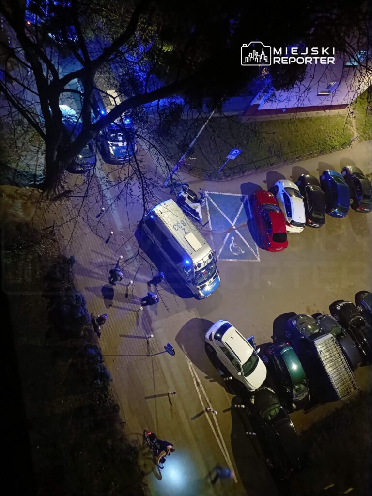 Nocna interwencja policji w oświetlonym niebieskim świetle, z radiowozem i grupą osób na chodniku.