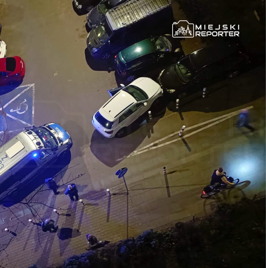 Grupa ludzi stoi obok zaparkowanego radiowozu z włączonymi światłami, na tle zatłoczonego parkingu nocą.