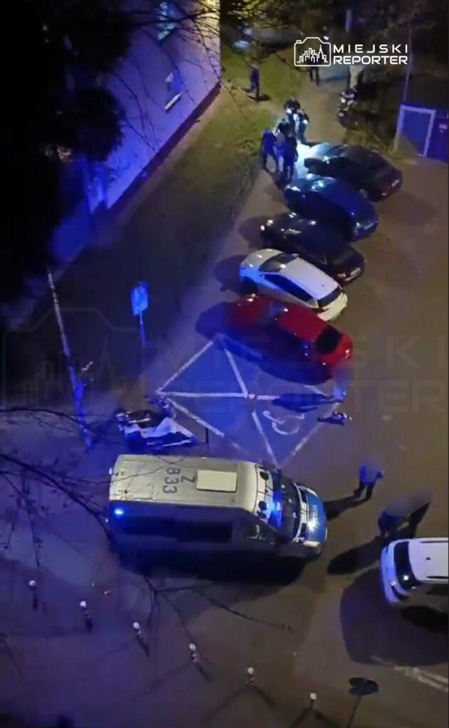 Grupa ludzi gromadzi się wokół radiowozu policyjnego na oświetlonym parkingu w nocy.