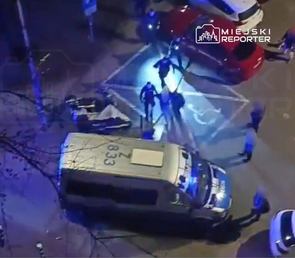 Grupa policjantów z latarkami przeszukuje ulicę w nocy, obok leży przewrócone auto.