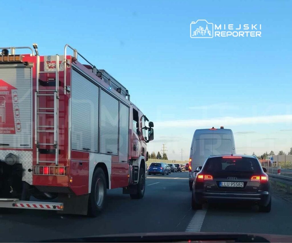 Czerwony wóz strażacki przejeżdża obok zatoru drogowego na autostradzie, w tle widoczne samochody osobowe.
