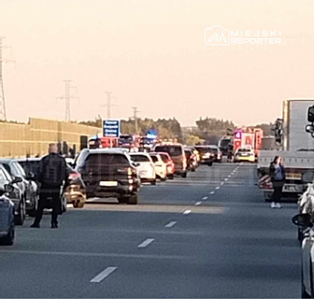 Kierowcy zatrzymani w korku na drodze, w tle widoczne pojazdy służb ratunkowych i policyjnych.
