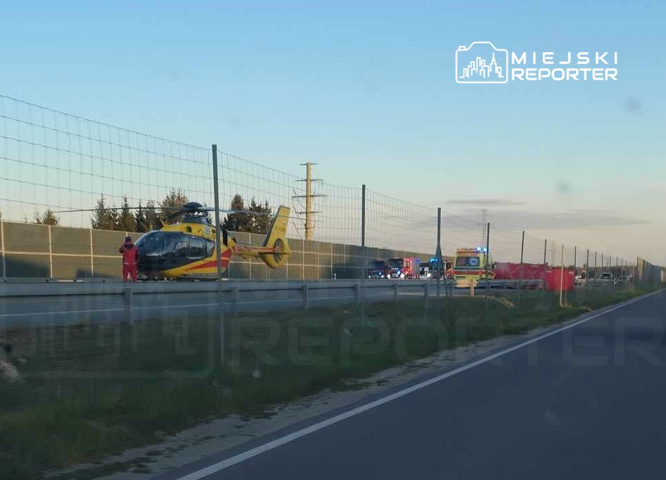Śmigłowiec ratunkowy lądował na poboczu drogi, obok stacji ambulansowej i pojazdów służb ratunkowych.