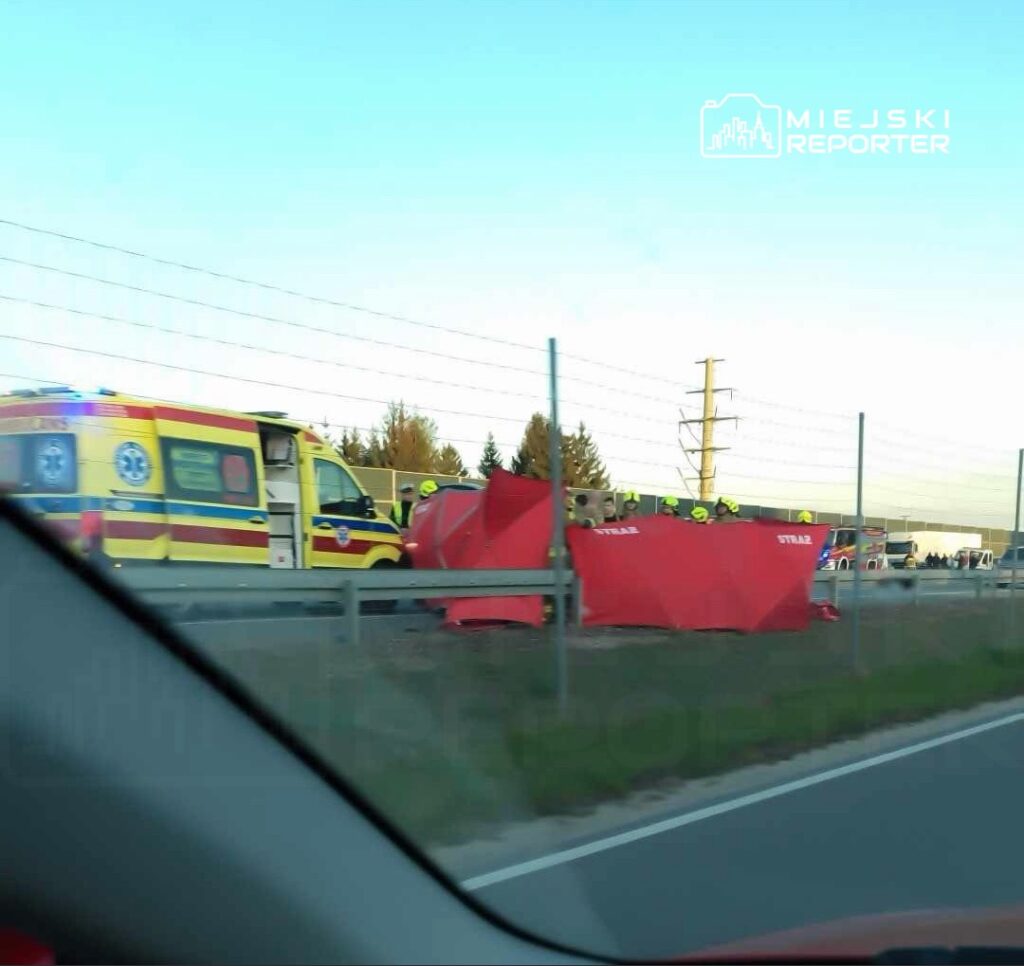Zespół ratunkowy pracuje przy wypadku drogowym, obok czerwonych namiotów zabezpieczających, na poboczu drogi.