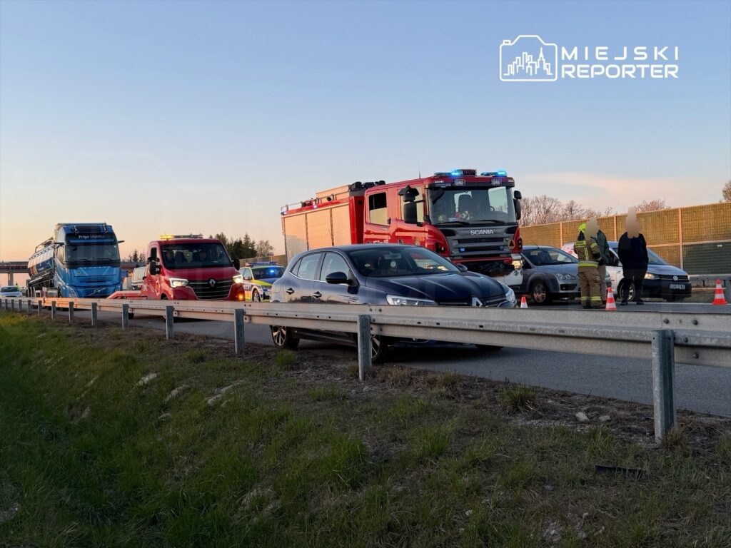Na drodze zatrzymane pojazdy, w tym niebieski autobus i czerwony wóz strażacki, a obok rozmawiający ratownicy.