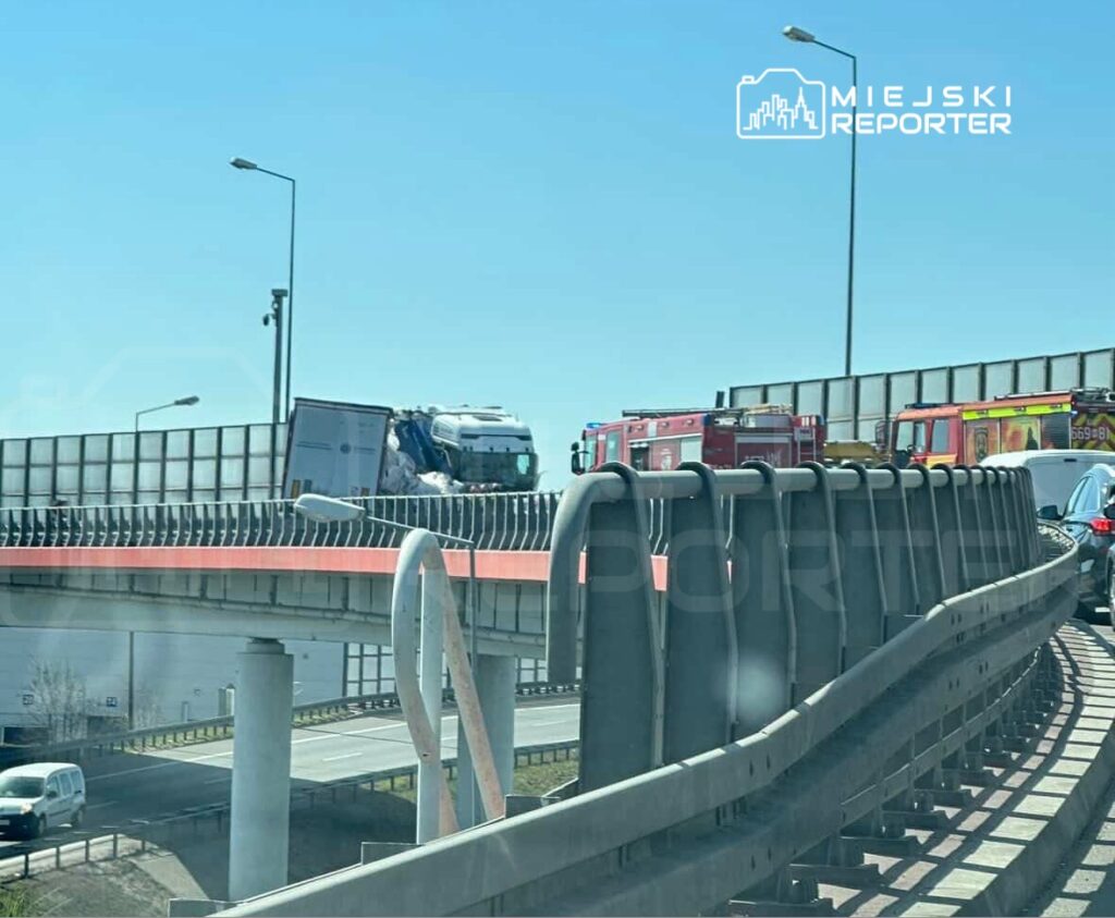 Ciągnik siodłowy z naczepą przewrócony na moście, obok stoją pojazdy straży pożarnej oraz zablokowane samochody osobowe.