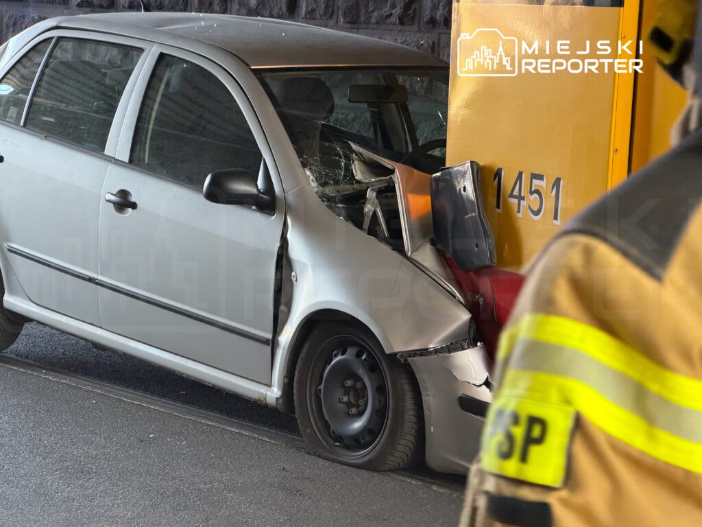 Uszkodzony srebrny samochód osobowy z wgniecioną tylną częścią, stojący obok żółtego pojazdu służb ratunkowych.