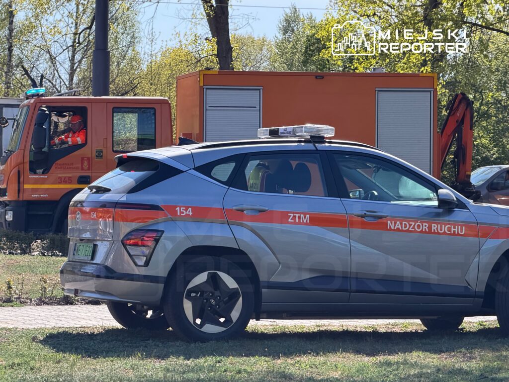 W tle widoczny pojazd służbowy z napisem "Nadzór Ruchu", a obok stoi samochód z niebieskim sygnalizatorem.