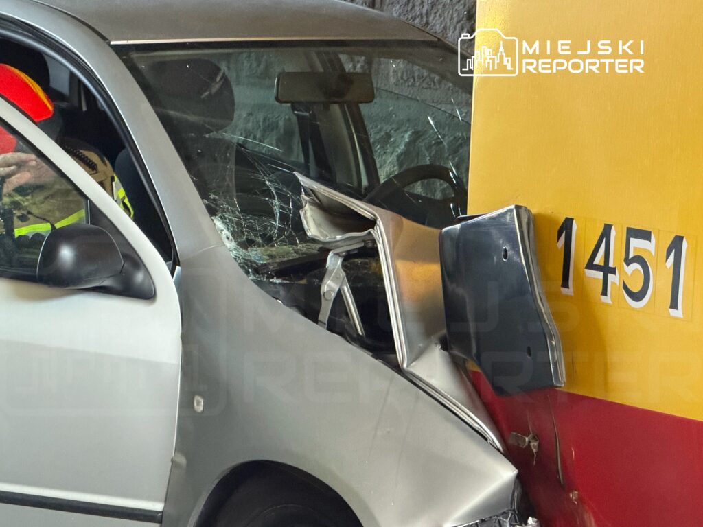 Zniszczony samochód osobowy uderzający w ścianę garażu, w tle widoczny ratownik w kamizelce odblaskowej.