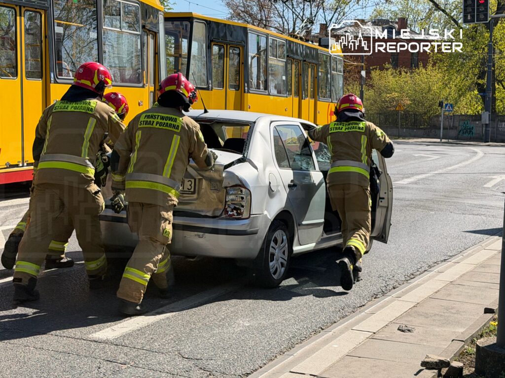 Czterech strażaków w mundurach z logo "Państwowa Straż Pożarna" otwiera drzwi samochodu na ulicy w pobliżu tramwaju.