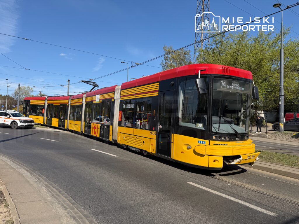 Tramwaj linii 74 zatrzymany na skrzyżowaniu, obok niego stoi radiowóz policyjny. W tle widoczna zieleń drzew.