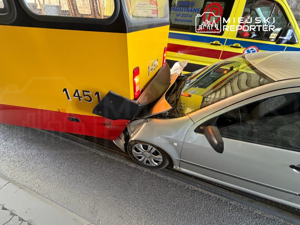 Złoty tramwaj z numerem 1451 zderza się z szarym samochodem osobowym na ulicy. W tle widać ambulans.