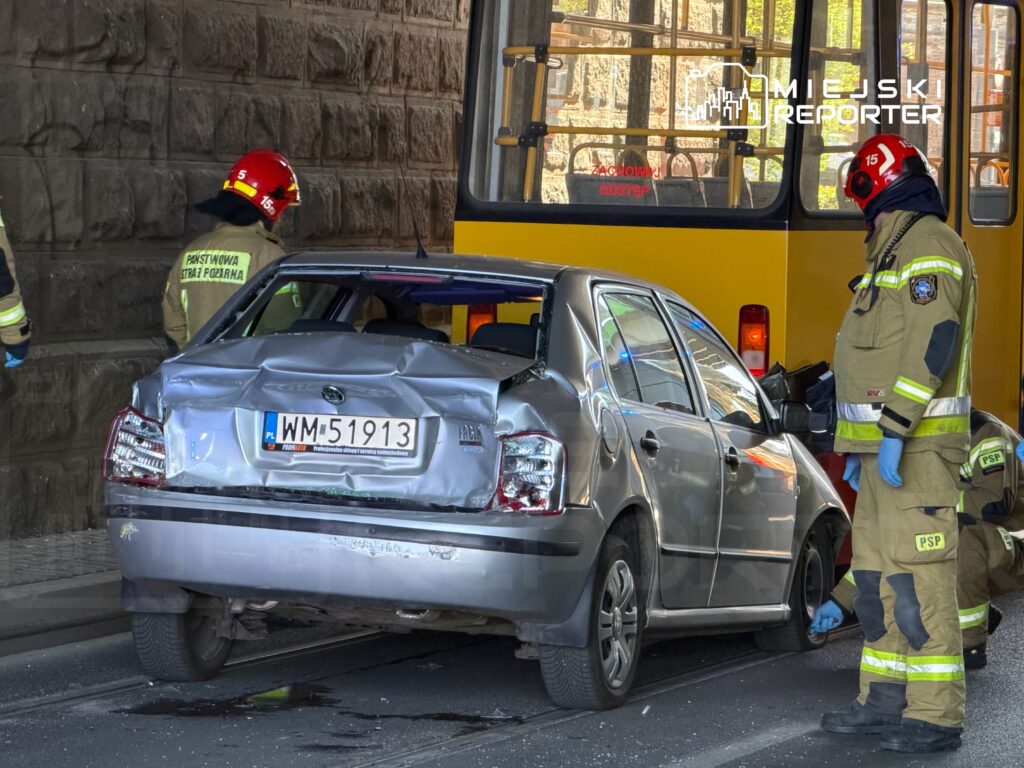 Samochód osobowy z uszkodzoną tylną częścią stoi na torach tramwajowych, obok pracują strażacy w mundurach.