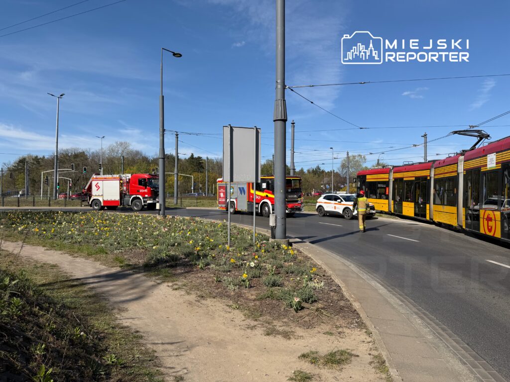 Straż pożarna i radiowóz policji na skrzyżowaniu, obok tramwajów, w otoczeniu kwitnących kwiatów.