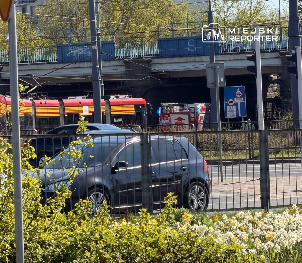Czarny samochód osobowy przejeżdża obok tramwaju oraz wozu strażackiego w tle, przy ruchliwej ulicy.