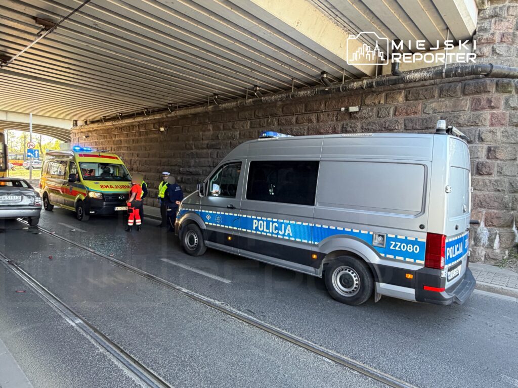 Karetka i radiowóz policyjny zatrzymane pod wiaduktem, w pobliżu uszkodzonego samochodu. Funkcjonariusze rozmawiają.