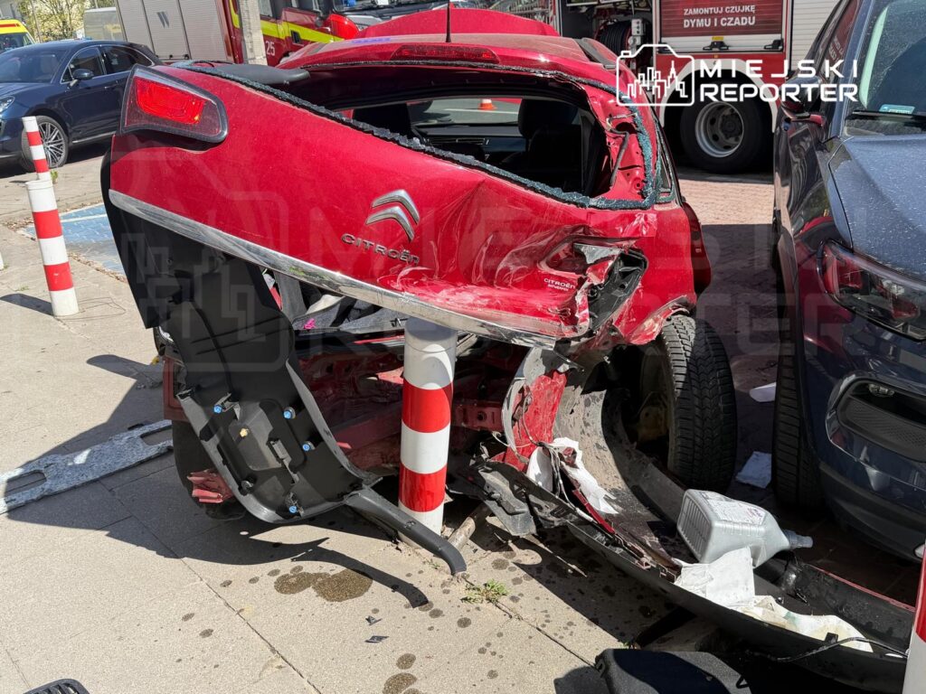 Czerwony samochód osobowy zniszczony w wyniku kolizji, zaparkowany obok stojaka na rowery, w tle inne pojazdy.