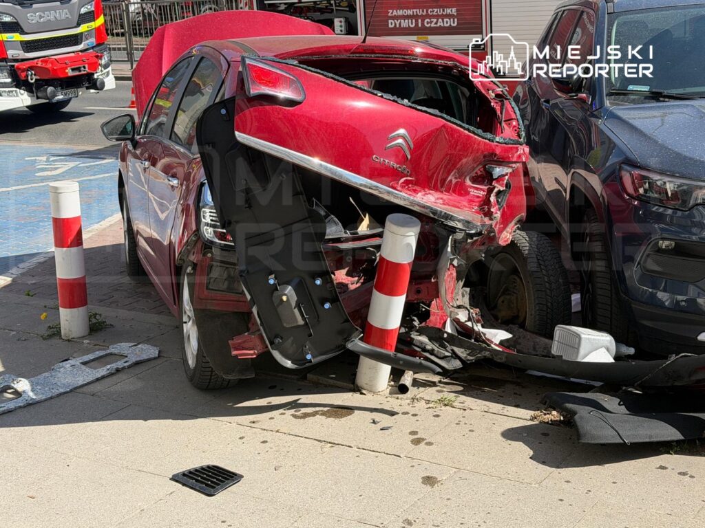 Czerwony samochód osobowy z uszkodzoną tylną częścią stoi na chodniku obok słupków ochronnych.