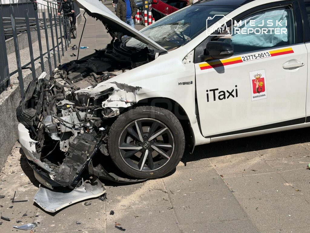 Uszkodzony samochód taxi z rozbitą przednią częścią stoi na chodniku, obok widać fragmenty zderzaka.