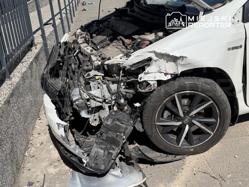 Uszkodzony samochód osobowy z oderwaną przednią częścią leży na chodniku obok metalowej balustrady.