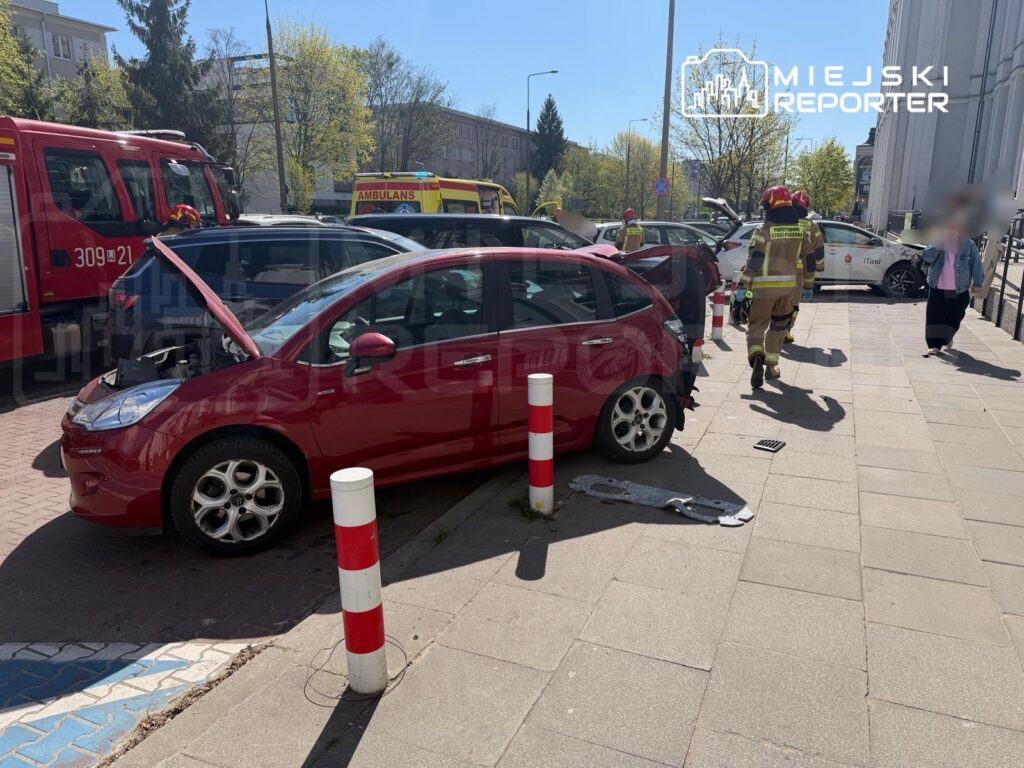 Czerwony samochód z otwartą maską stoi na chodniku, obok strażacy w mundurach podchodzą do zdarzenia.
