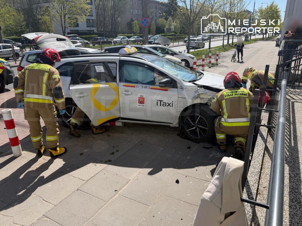 Strażacy w mundurach pracują nad usunięciem uszkodzonego samochodu osobowego na chodniku w miejskim otoczeniu.