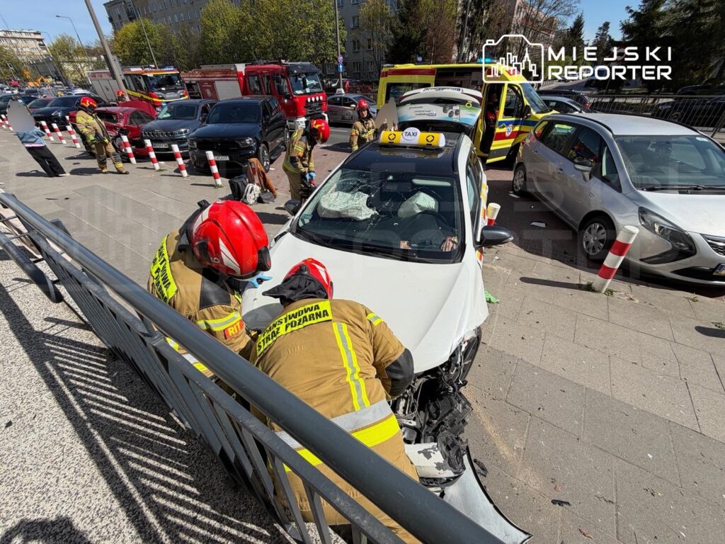 Dwaj strażacy w hełmach pracują nad usunięciem zniszczonego pojazdu w miejskim otoczeniu, obok innych samochodów.